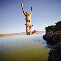 Young woman jumping off edge of cliff into river with arms overhead