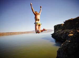 Young woman jumping off edge of cliff into river with arms overhead