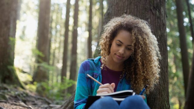 Woman writing in journal against tree in woods