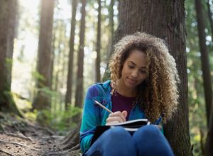 Woman writing in journal against tree in woods