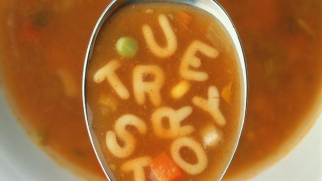 Spoon over bowl of alphabet soup, letters spelling 'true story'