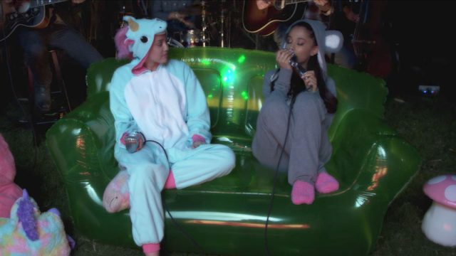 Ariana Grande and Miley Cyrus wearing onesies singing on a green sofa.