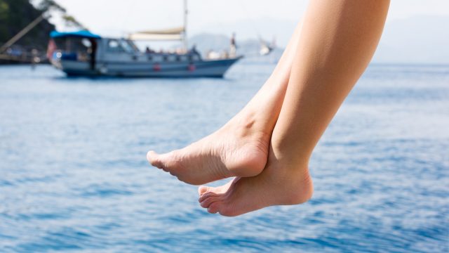 Legs dangling off a dock over the ocean with a boat in the background.