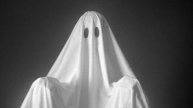 Person Wearing A Ghost Costume, Made Out Of A White Sheet With Two Holes In It. Highlights Are On The Sheet, The Background Is Pitch Black.