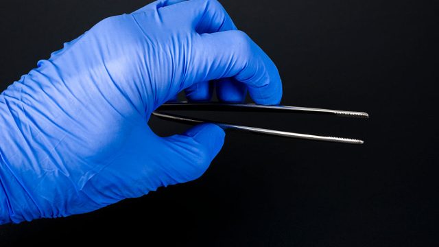 A hand in a blue medical glove is holding a tweezers for medical use, displayed on a black table.