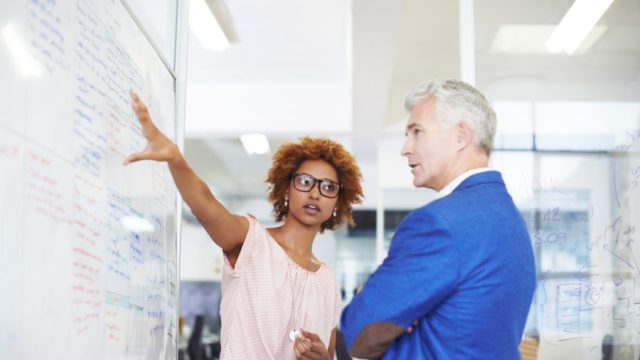 Young businesswoman explaining strategy on whiteboard to male colleague in creative workspace