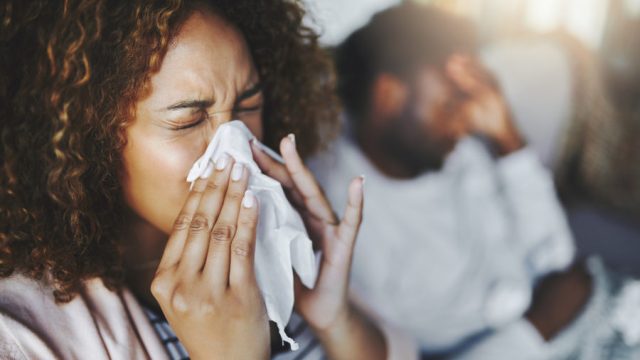 Shot of a young woman blowing her nose with her boyfriend in the background