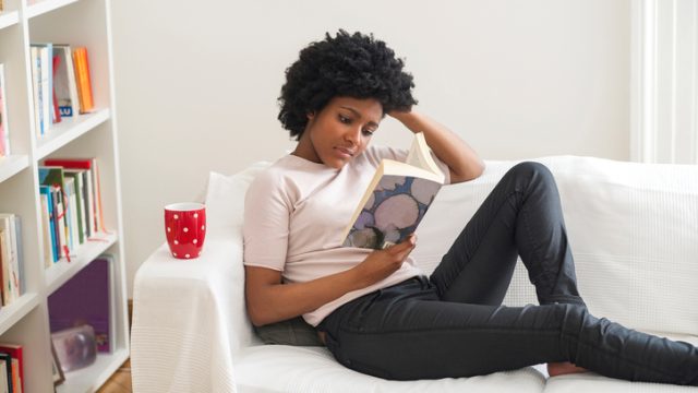 Black woman reading book on sofa