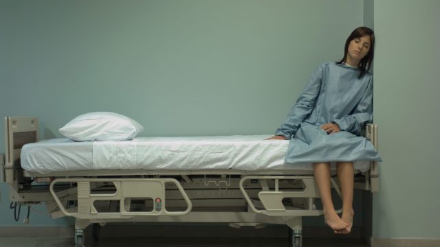 Female patient sitting on hospital bed with eyes closed, leaning head against wall