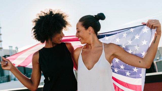 Female friends holding US American flag, standing on rooftop