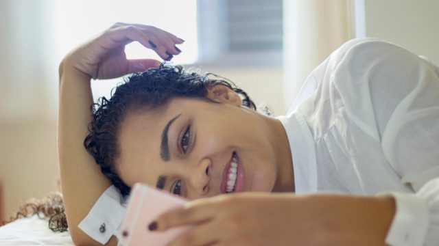 Happy woman sitting on bed reading text message on smartphone. Smiling girl sitting in bedroom in casual in conversation with friend in a chat room.