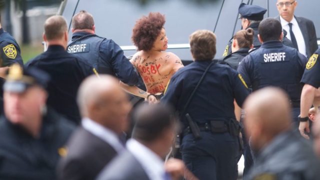 A topless protester lunged at Bill Cosby outside the courthouse where he faces a retrial.