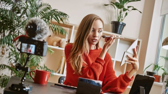 Makeup tutorial. Cute and young blogger using brush to apply highlighter while broadcasting live video to social network