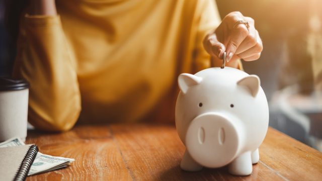 Woman Putting Coin In Piggy Bank On Table At Home